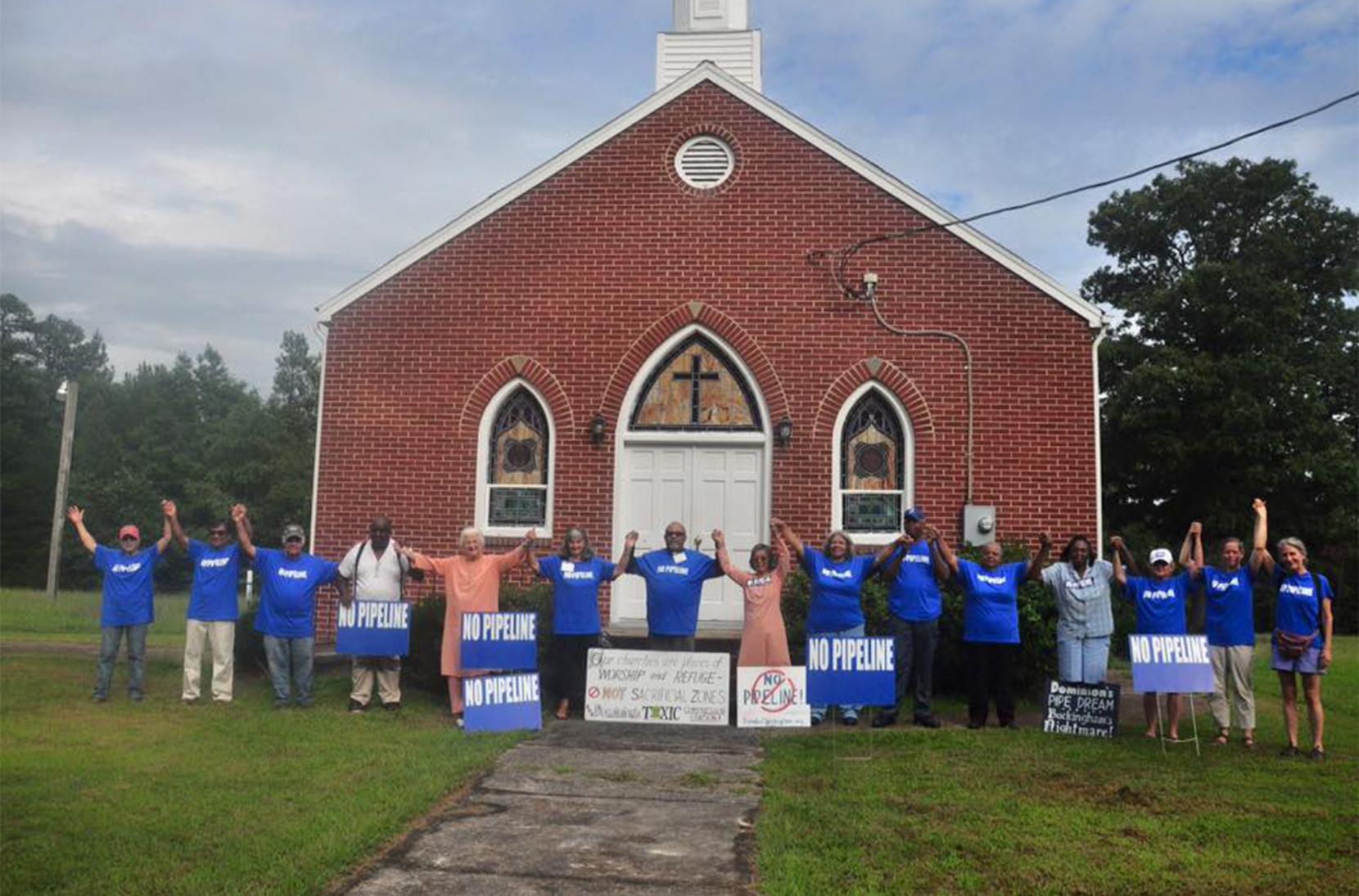 Fifteen community activists, mostly in matching No Pipeline T-shirts, joyfully hold hands in front of a small brick church in celebration of the cancellation of the Atlantic Coast Pipeline. Several anti-pipeline protest signs lean up against them.
