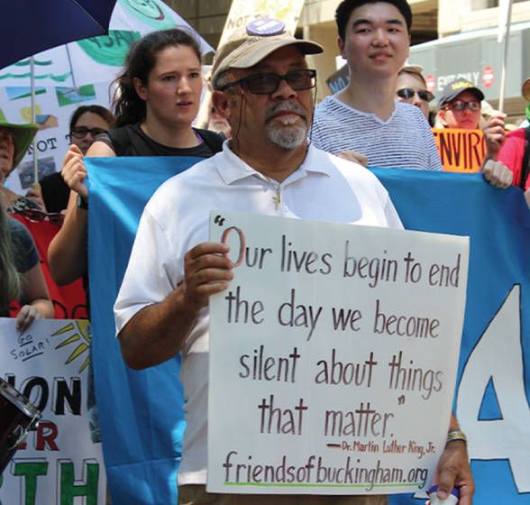 A man in a beige baseball cap and white button-up holds a sign that reads "Our lives begin to end the day we become silent about things that matter" at a Buckingham protest.