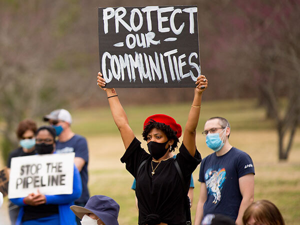 A protestor in a black medical mask and a red hat holds up a sign that reads "Protect our Communities".