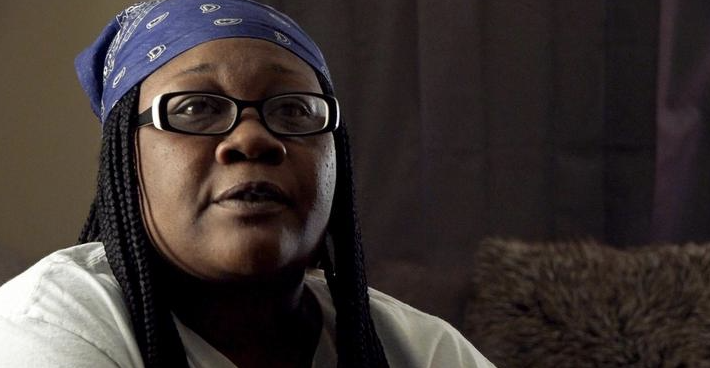 A woman in high contrast with oval glasses, braids, and a blue bandana speaks to the camera.
