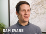 Sam Evans smiles in a collared gray sweater in front of a textured topographical map.