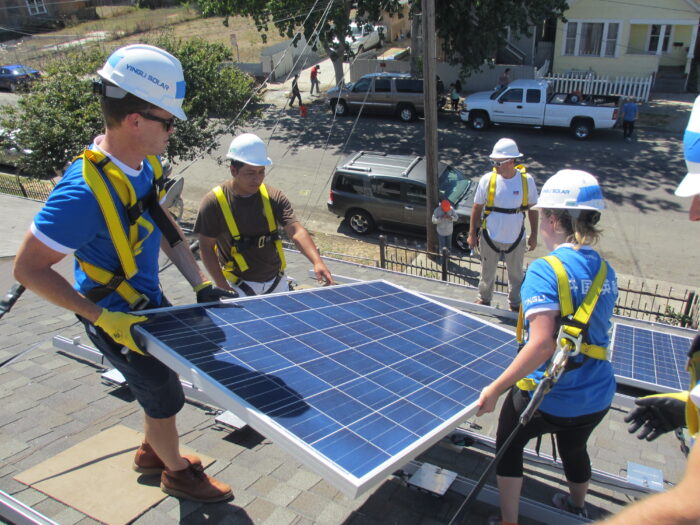 Three workers in hard hats and safety gear install a rooftop solar panel while a supervisor guides them.