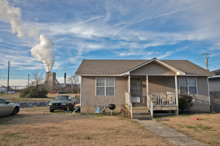 A car is parked beside a tan one story home with a small front porch as large smokestacks release plumes of smoke in the background.