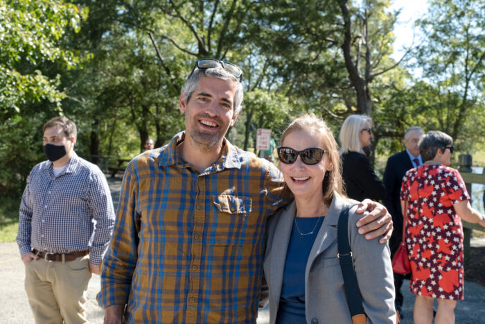 A man in a plaid shirt and woman in business wear stand outside in a small crowd and pose for the camera.