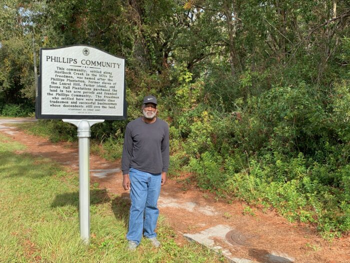 A tall man in jeans, sweatshirt and baseball cap stands next to a historical marker on the side of the road with information about the Phillips Community outside of Charleston, South Carolina.