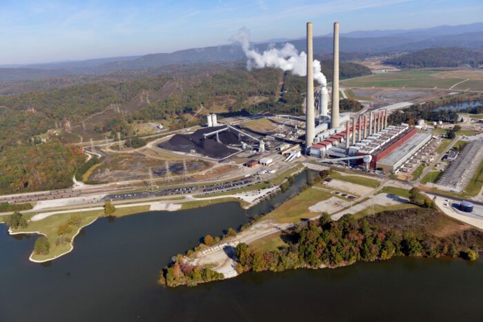 An aerial view shows a river in the foreground and a large power plant with smokestacks on its shores.