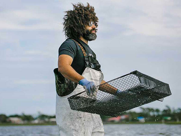 A man in waders, a tshirt and sunglasses carries a shallow wire basket with work gloves in knee-high water.