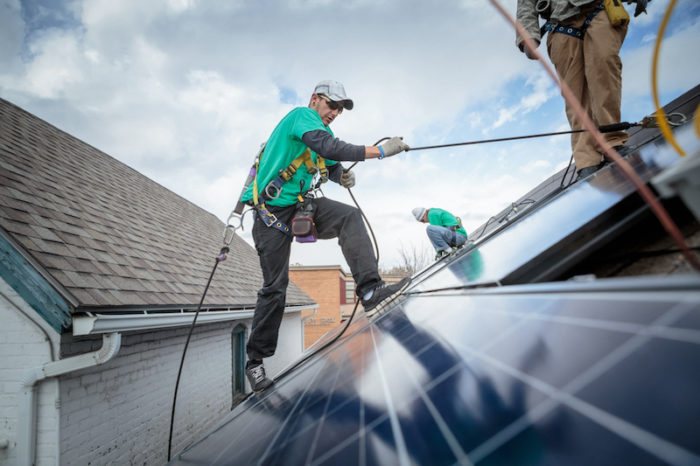 Man in a green shirt scales the side of a roof covered in solar panels.