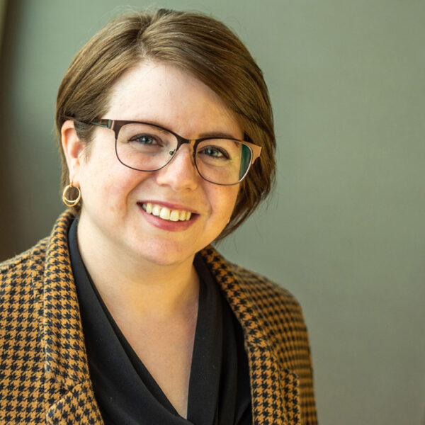 Portrait of Catherine Crafa smiling in an orange houndstooth patterned blazer, gold earrings, and wire-rimmed glasses.
