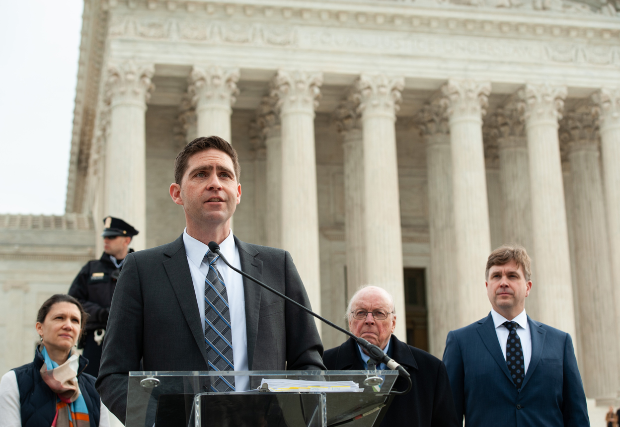 A man stands at a podium in front of the U.S. Supreme Court with clients and colleagues lined up behind him.