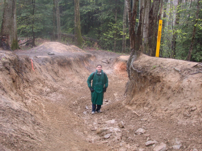 A man wearing green rain gear stands in a long muddy ditch taller than him on both sides, looking up a steep hill with trees on either side of the ditch.