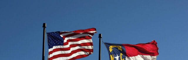 The flag of the United States and the flag of North Carolina fly side-by-side against a blue sky.