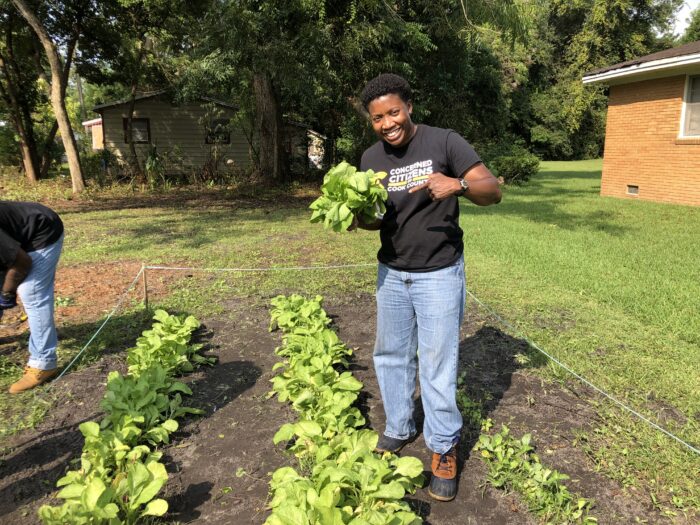 Dr. Treva Gear stands in a garden bed of lettuce. In a black Concerned Citizens of Cook County t-shirt and loose blue jeans, she smiles while pointing to the lush head of lettuce she's holding.
