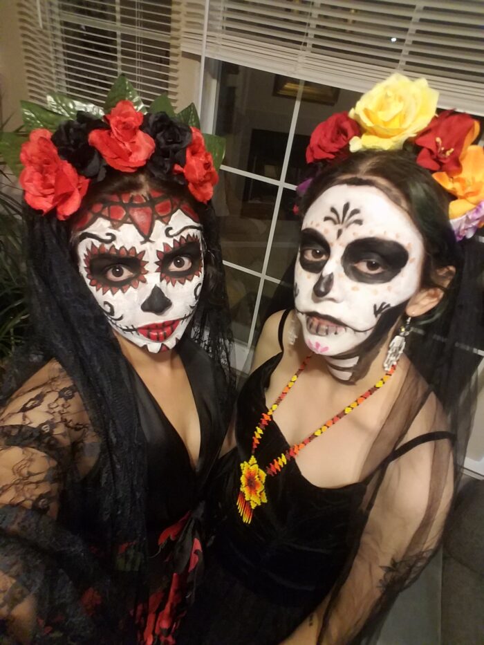 Two Mexican women look into the camera with flower crowns and faces painted as skeletons for Dia de los Muertos.