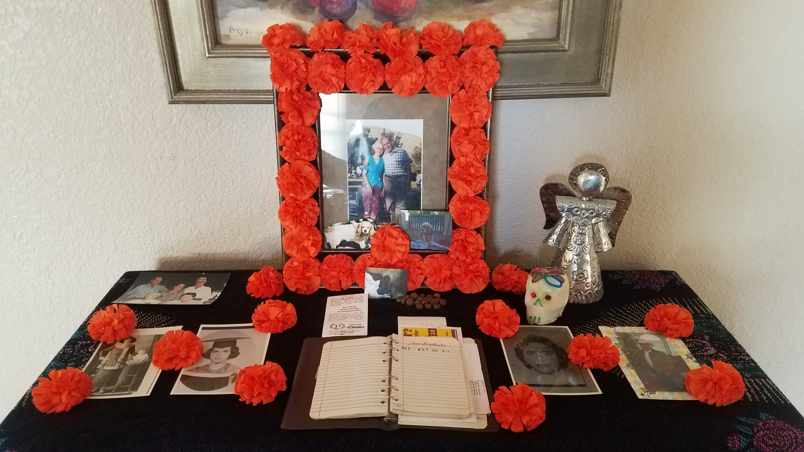 An altar featuring red flowers, photographs of loved ones and small keepsakes including a sugar skull and an angel figurine.
