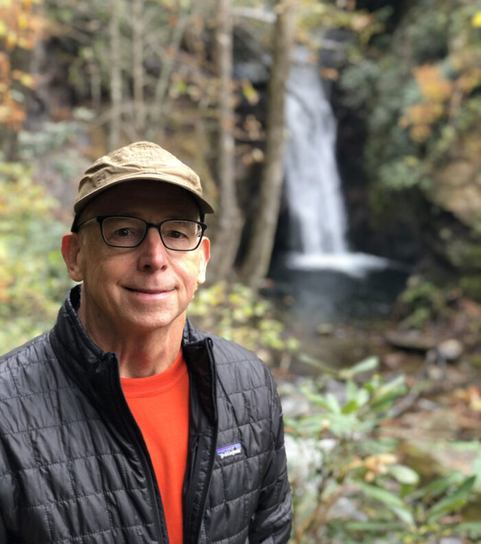 Man on a hike poses in front of a waterfall wearing a ball cap and thin down coat over a tshirt