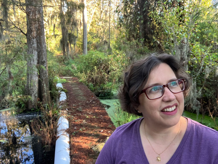 Woman in a purple tshirt and glasses looks up while on a boardwalk trail with a lush swamp in the background.
