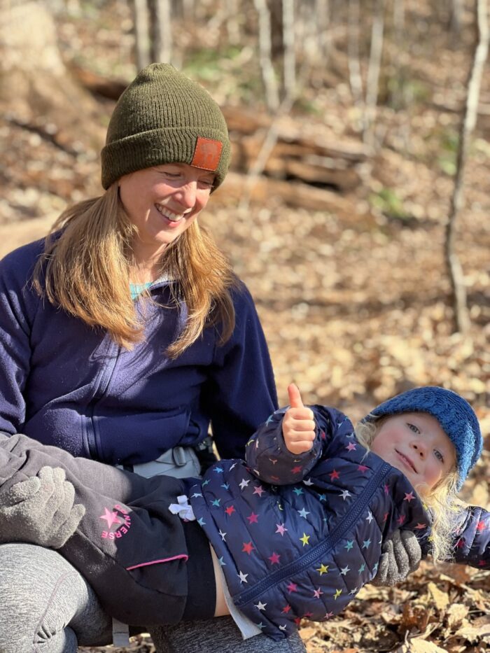 Ramona McGee holds her daughter, who is playfully giving a thumbs up, out on a wooded trail. Both are bundled up in winter gear including hats, coats, and gloves.