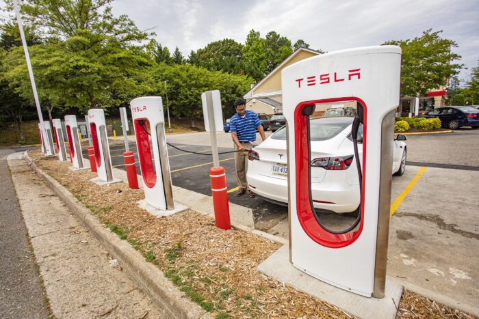 A man charges a white electric car at a row of Tesla-brand electric car chargers.