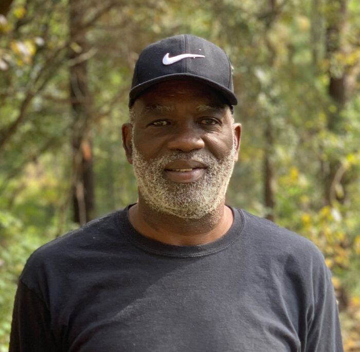 Black man with a gray beard stands in front of a wooded area wearing a black long sleeve tee and a black Nike baseball hat.