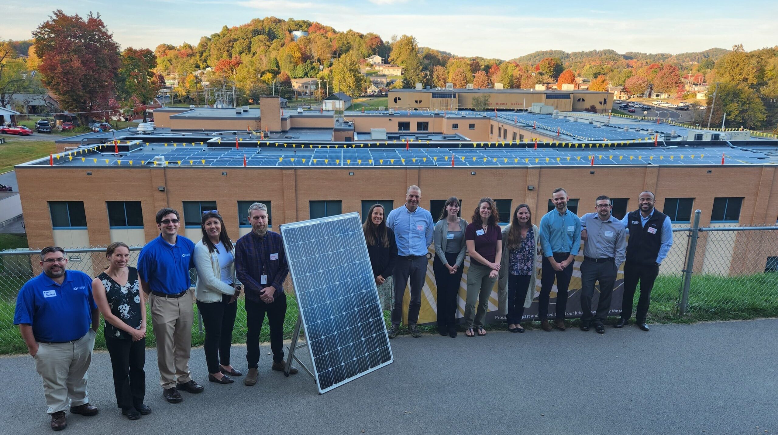 A line of people stand on a sidewalk with a demo solar panel. Behind them is a school roof covered in solar panels and a tree-covered mountain in the background.