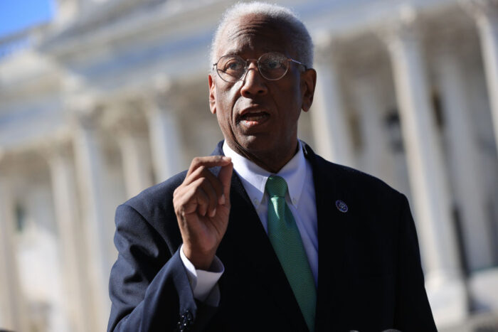 A Black man in a navy suit, white shirt and green tie stands in front of the columns of a government building.