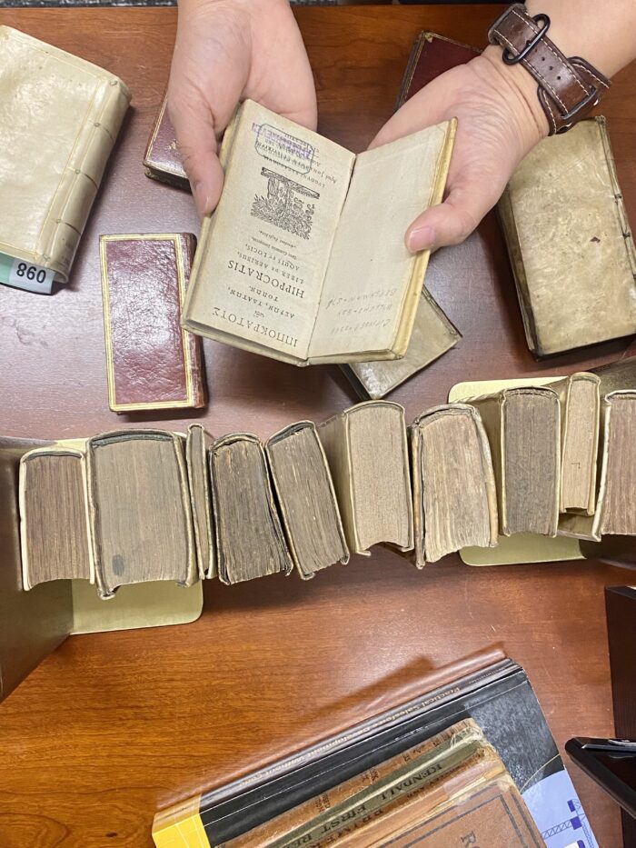 An overhead shot shows old books lined up on Donna Sy's desk. She holds an Elzevier-printed Hippocrates book called, "Airs, Waters, Places," which describes ancient theories of how the environment affects the physical and mental health of the people born and raised there.