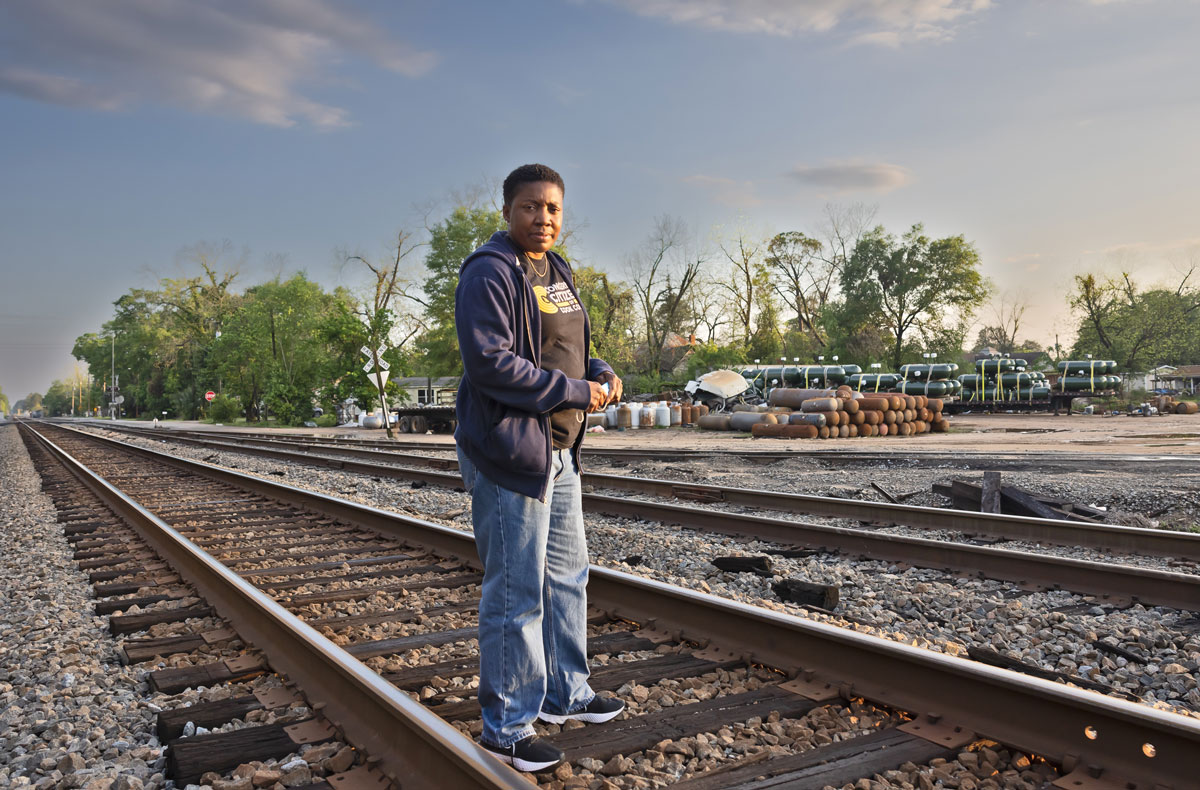 A Black woman with short hair in jeans and Black hoodie stands in the railroad tracks.