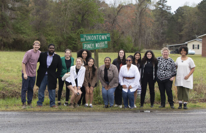 A group of about a dozen people pose in front of a rural street sign at the intersection of Uniontown Road and Piney Woods Road.