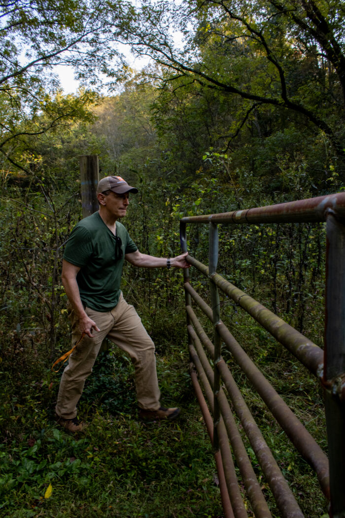 A farmer in dark work clothes and a ball cap swings open a rusted cattle gate.