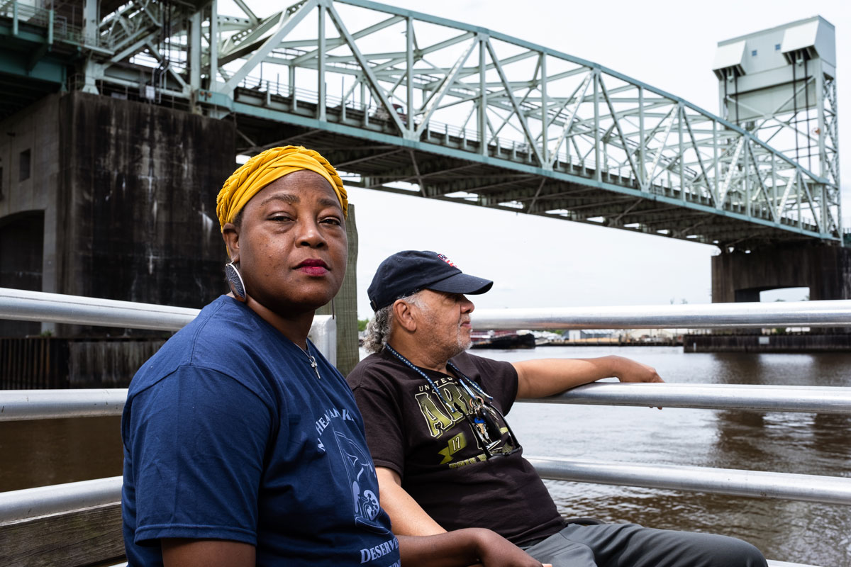 A woman in a yellow hair wrap and a man with gray hair under a black ball cap sit, holding hands, along a large river, with a bridge in the background.