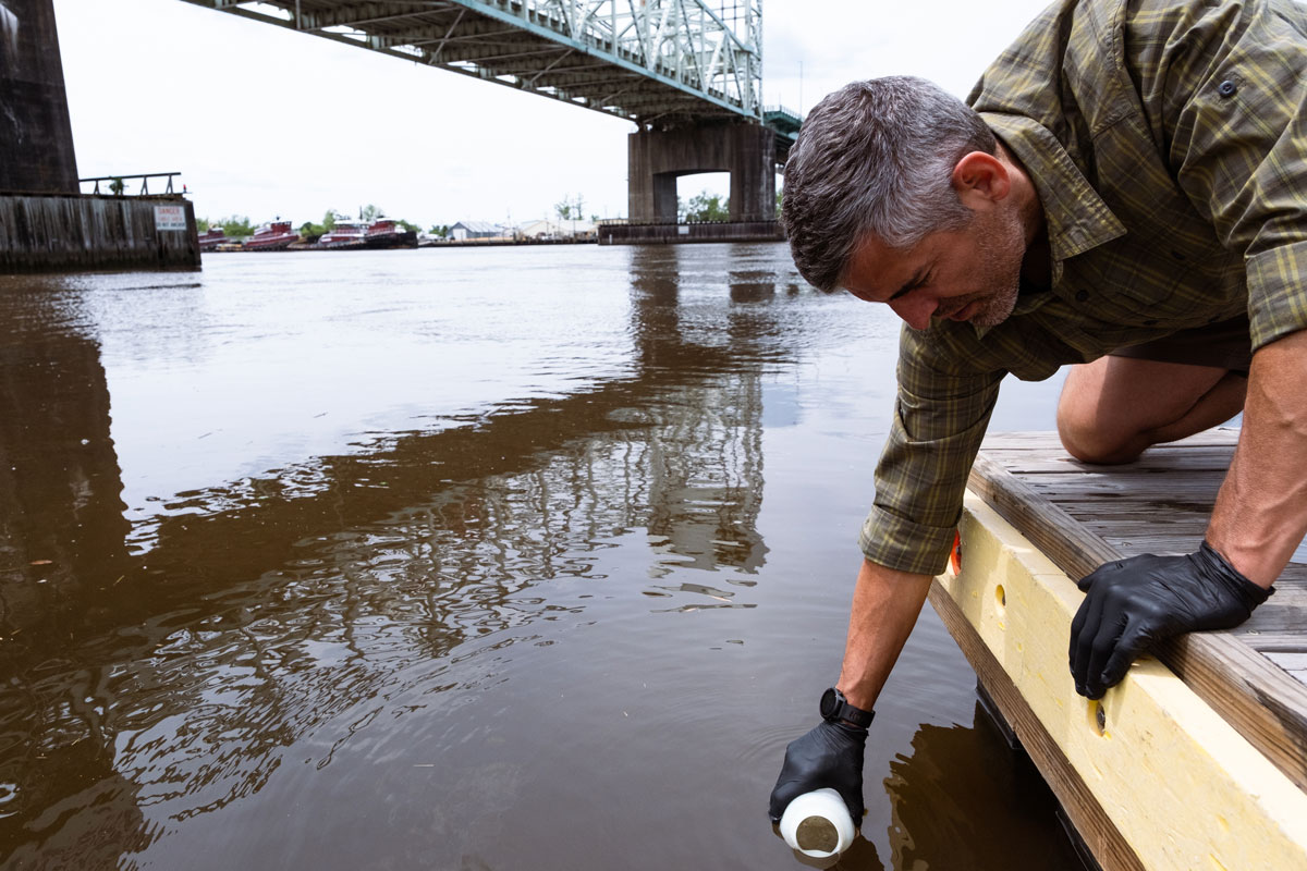 A man wearing gloves leans off a low dock to scoop dark river water into a plastic sampling jar.