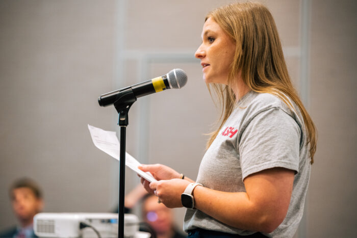 The profile of a young, blonde-haired woman in a grey t-shirt speaking into a microphone.